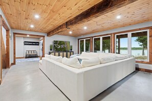 A spacious living area with a large white sofa, wooden ceiling, and a view of a lake through large windows. - Redstart Retreat | Hot Tub | Sauna | Waterfront | EV | (Miller Lake)