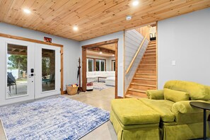 Een kamer met een houten plafond, een geel sofa, een vloerkleed met blauw patroon en een trap naar een andere kamer.