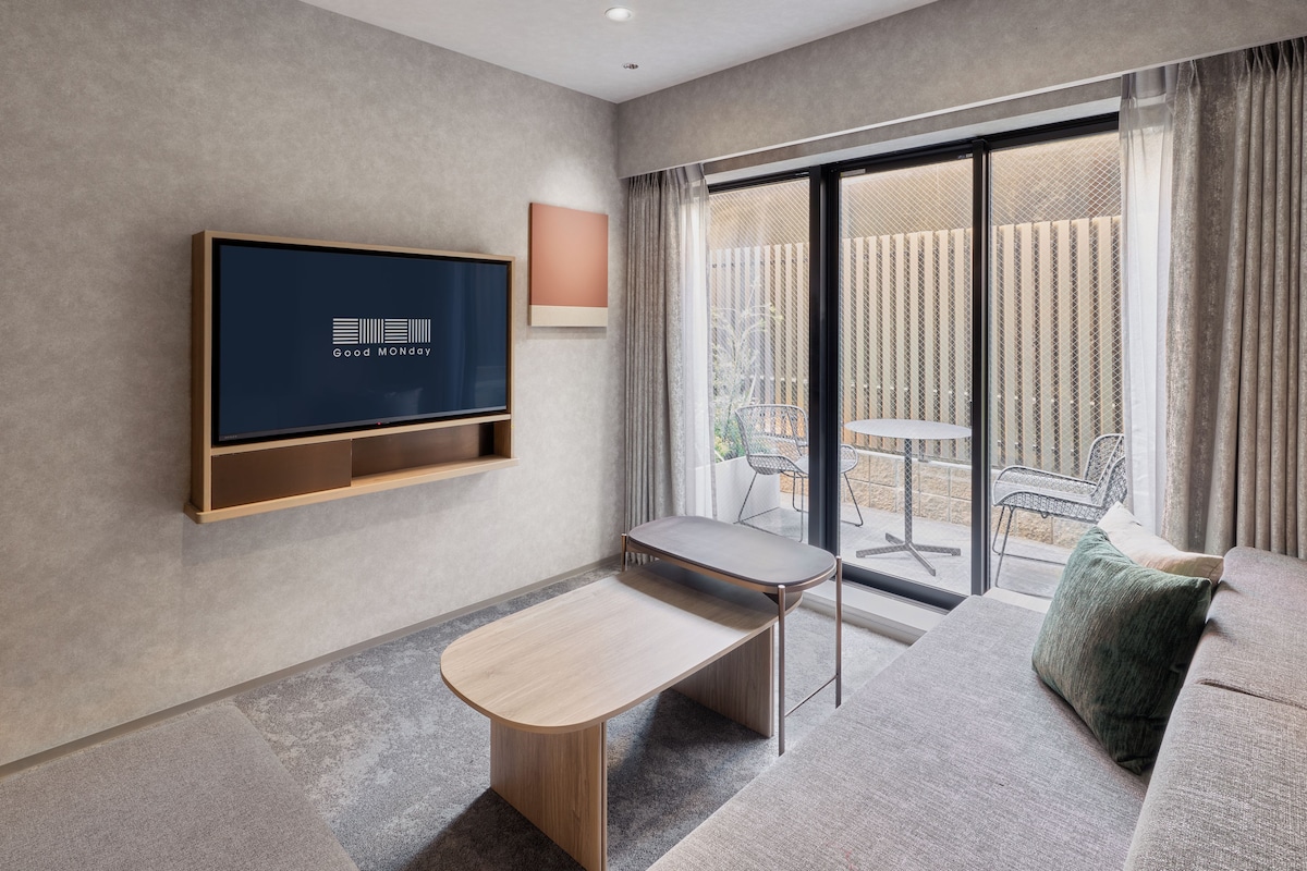 A modern hotel room with a flat-screen TV, a small wooden table, a sofa, and a sliding glass door leading to an outdoor area.