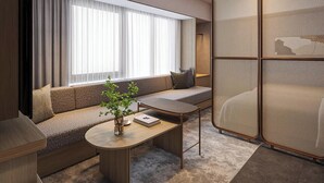 A modern hotel room with a sofa, a small table, a potted plant, and a large window with curtains.