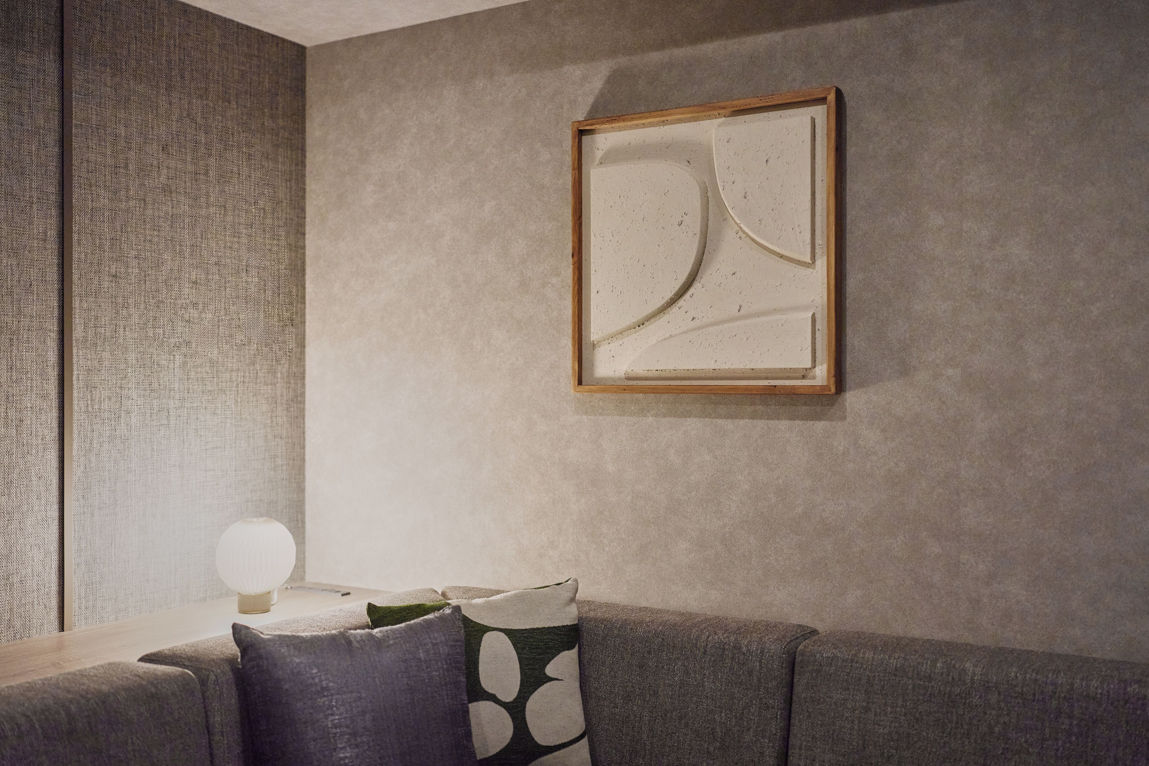 A living room with a textured wall, a framed abstract artwork, a round table with a lamp, and a sofa with patterned cushions.