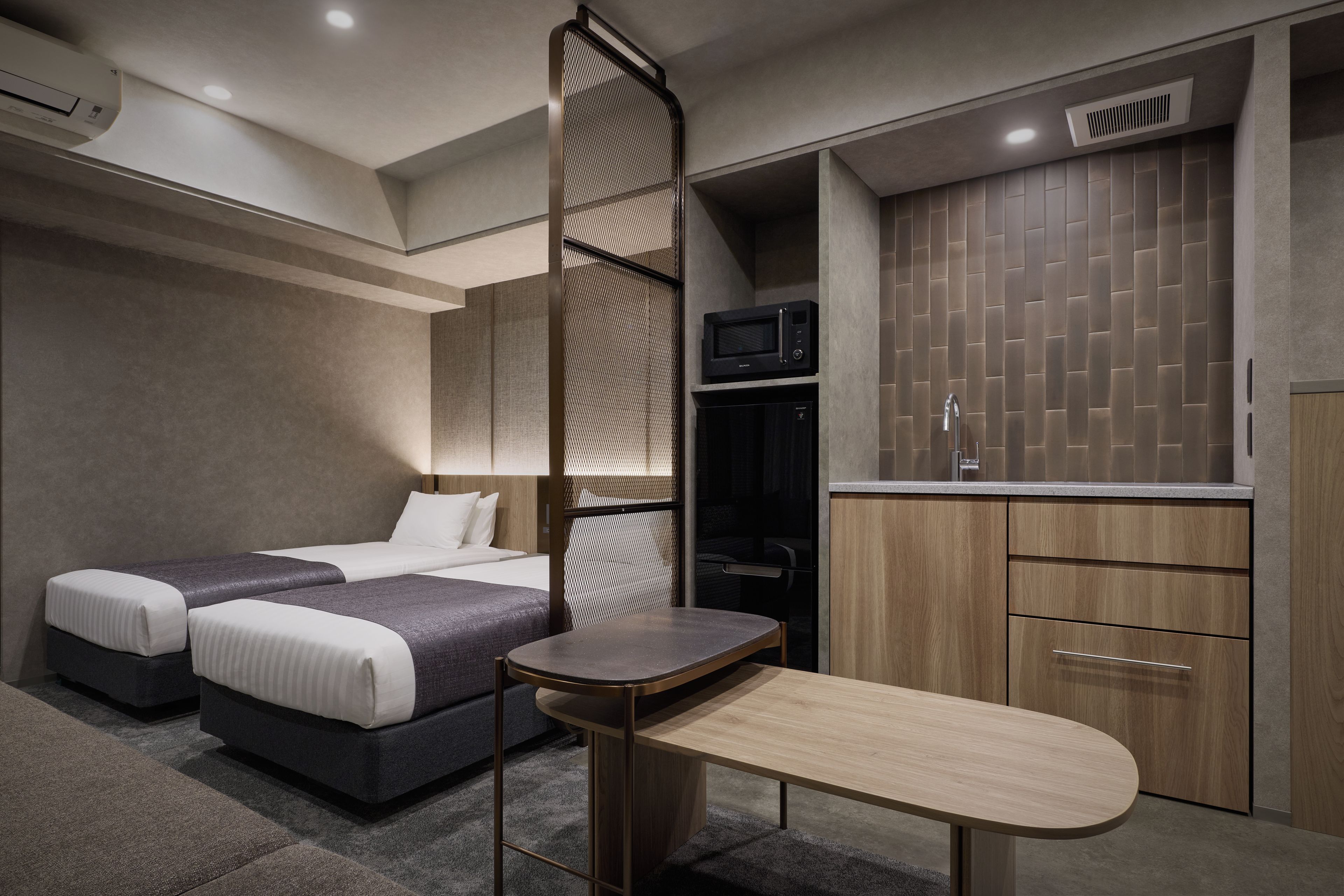 A modern hotel room with two beds, a small table, a kitchenette, and a wardrobe.