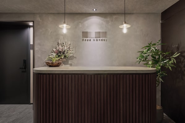 A reception desk with a modern design, featuring a wooden panel back and a grey countertop. There are two pendant lights above the desk, and a decorative plant is placed on the left side. - Good MONday ASAKUSA (Tokyo)