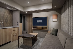 A modern living room with a flat-screen TV mounted on the wall, a sofa, a small table, and a kitchen area with wooden cabinets and a sink. - Good MONday ASAKUSA (Tokyo)