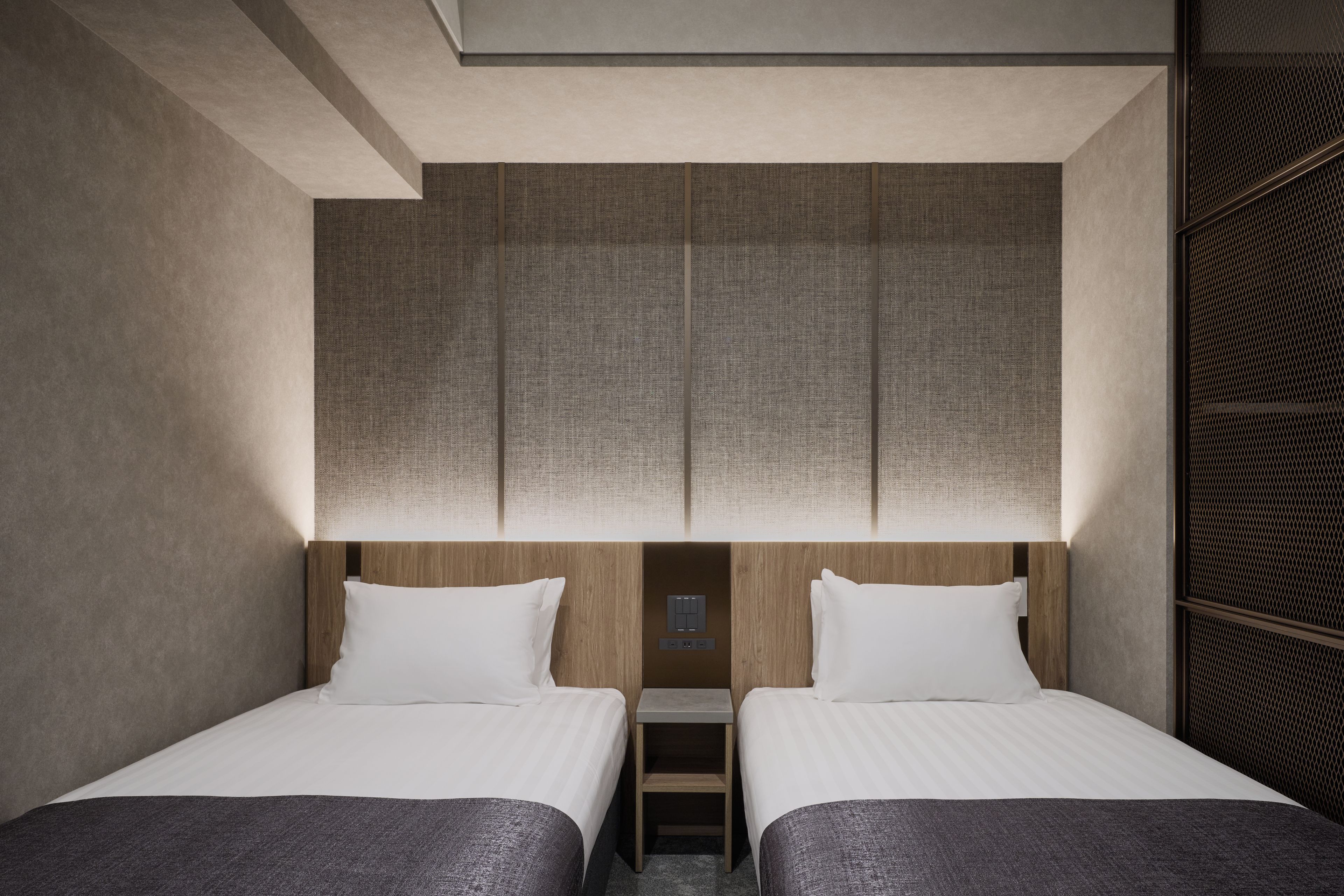 A hotel room with two beds, a wooden headboard, a small bedside table, and a wall-mounted control panel.