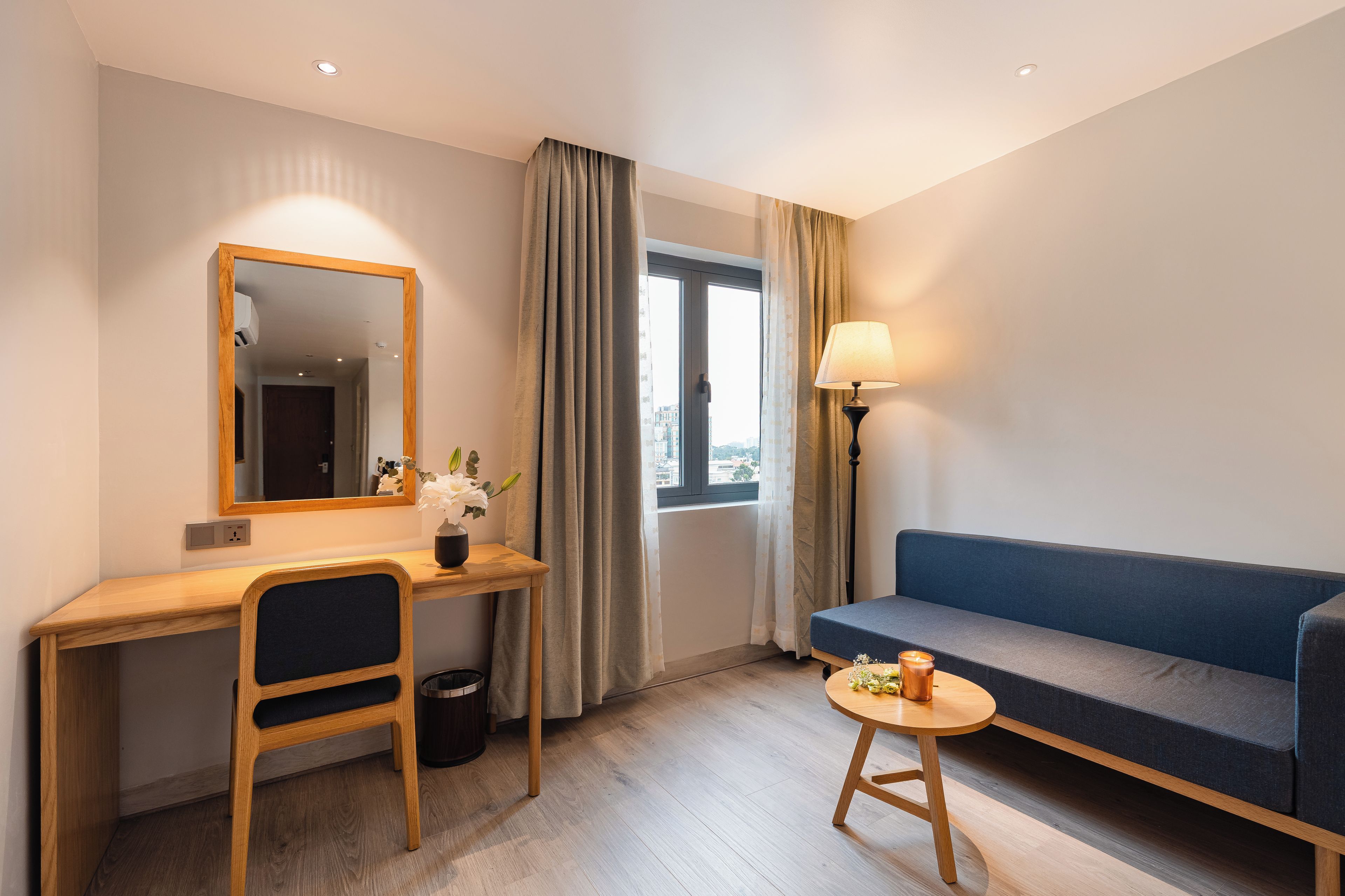 Habitación de hotel con un escritorio de madera, una silla, un sofá, una mesita redonda con lámpara y una ventana con cortinas.