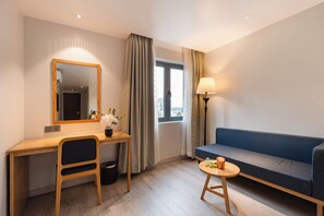 Une chambre d’hôtel avec un bureau en bois, une chaise, un canapé, une petite table ronde avec une lampe et une fenêtre avec des rideaux.