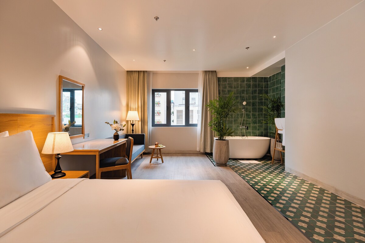 Et hotelværelse med seng, skrivebord, stol, et vindue, badeværelse med badekar og en potteplante.