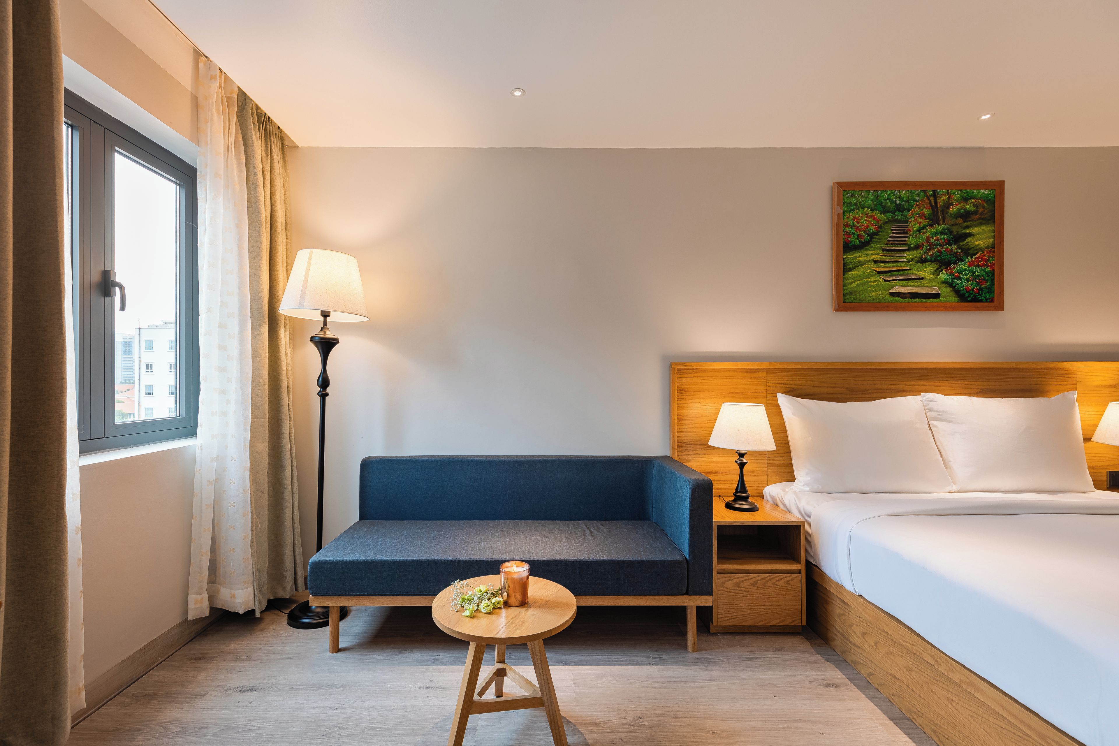 Habitación de hotel con cama, escritorio, silla, una pequeña mesa con una vela y un cuadro en la pared.