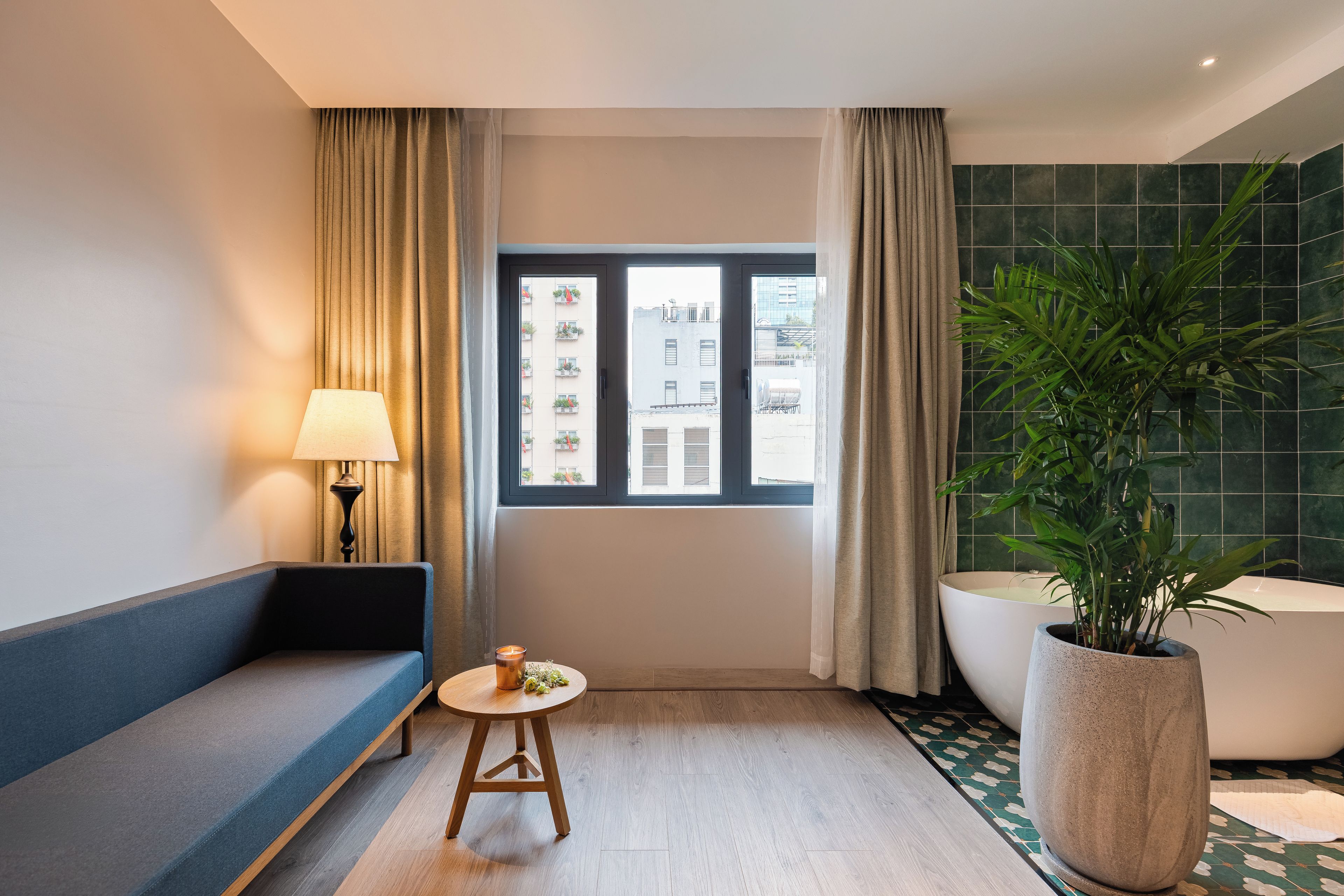 Habitación de hotel con sofá, una mesita de noche con lámpara, ventana con cortinas, una planta grande en maceta y una bañera.