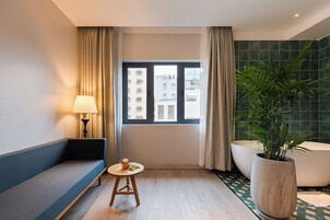 A hotel room with a sofa, a small table with a lamp, a window with curtains, a large potted plant, and a bathtub.