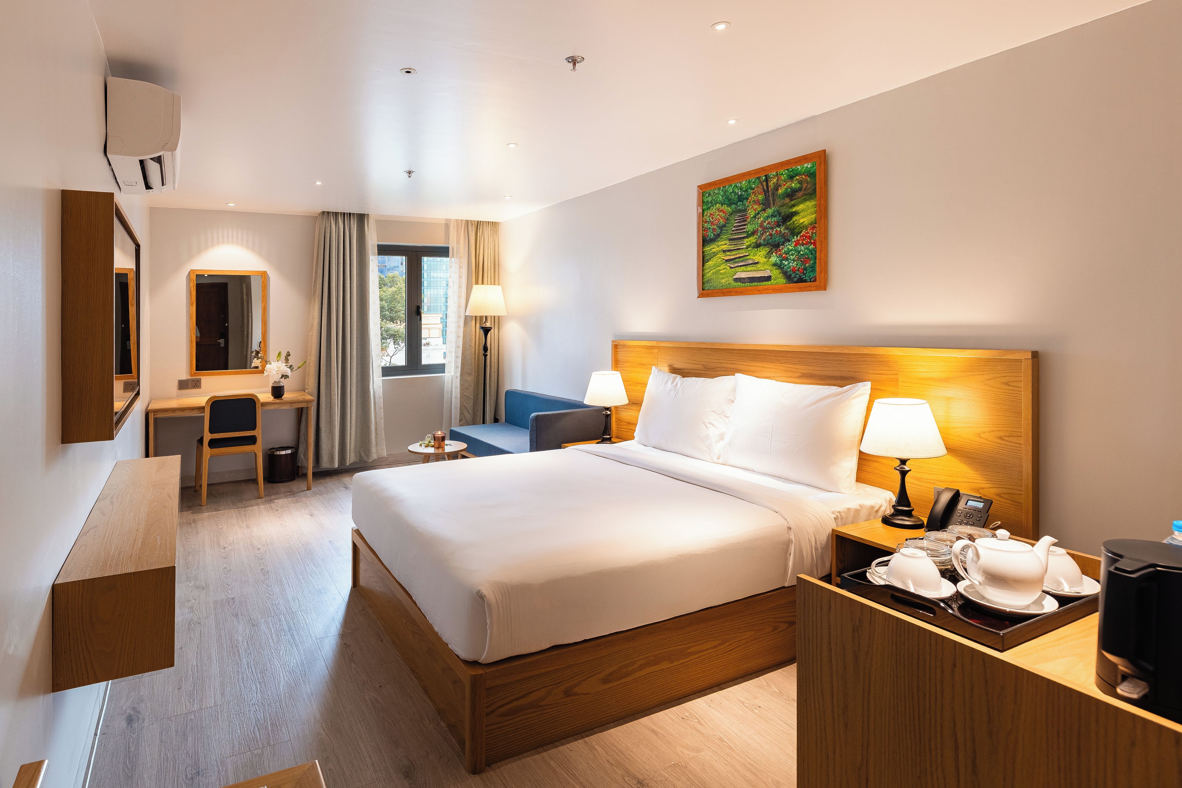 A hotel room with a large bed, a desk, a chair, a bedside table with a lamp, and a painting on the wall.