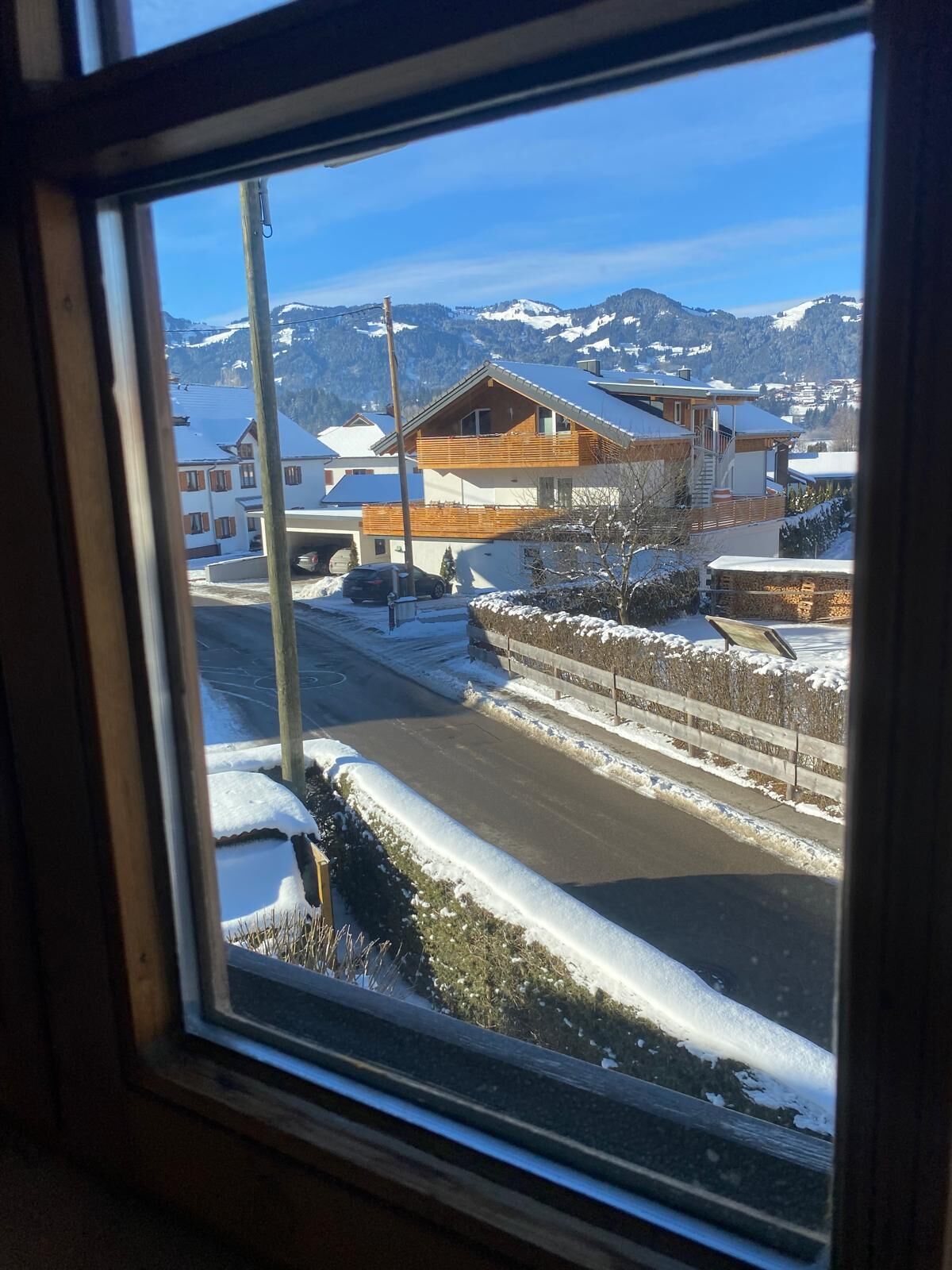 Blick aus dem Fenster auf eine Wohngegend mit Häusern, einer Straße und verschneiten Bergen im Hintergrund.