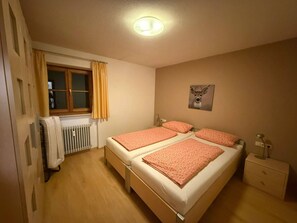Ein Schlafzimmer mit Doppelbett, Heizkörper, einem fenster mit Vorhängen, einer Wanduhr und einem kleinen Nachttisch.