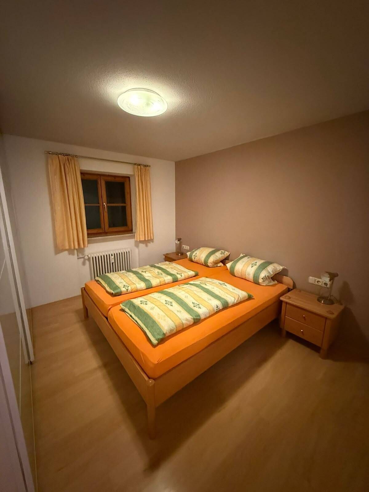 Ein Einzelzimmer mit einem hölzernen Bett, einem Nachttisch, einem Heizkörper und einem Fenster mit Vorhängen.