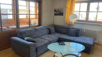 Ein modernes Wohnzimmer mit einem Ecksofa, einem Glastisch und Blick ins Freie durch große Fenster.