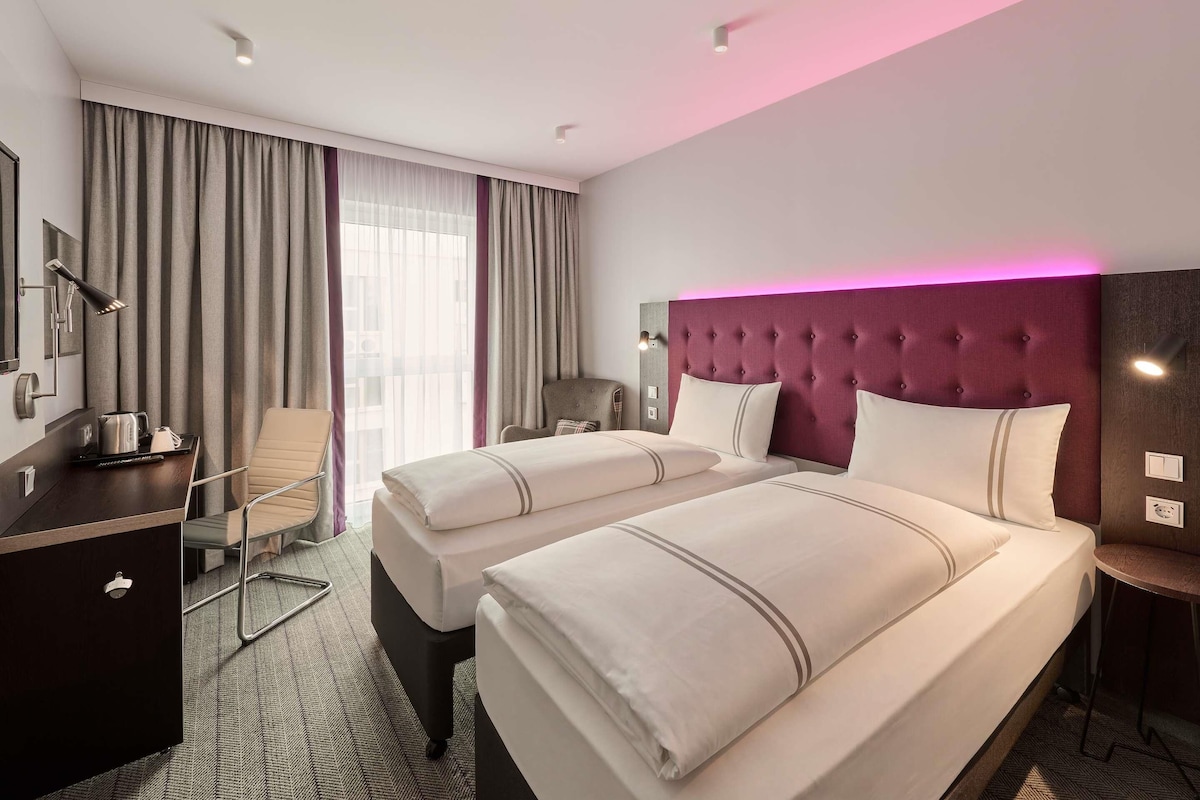 A hotel room with two beds, a desk, a chair, a desk lamp, and a window with curtains.