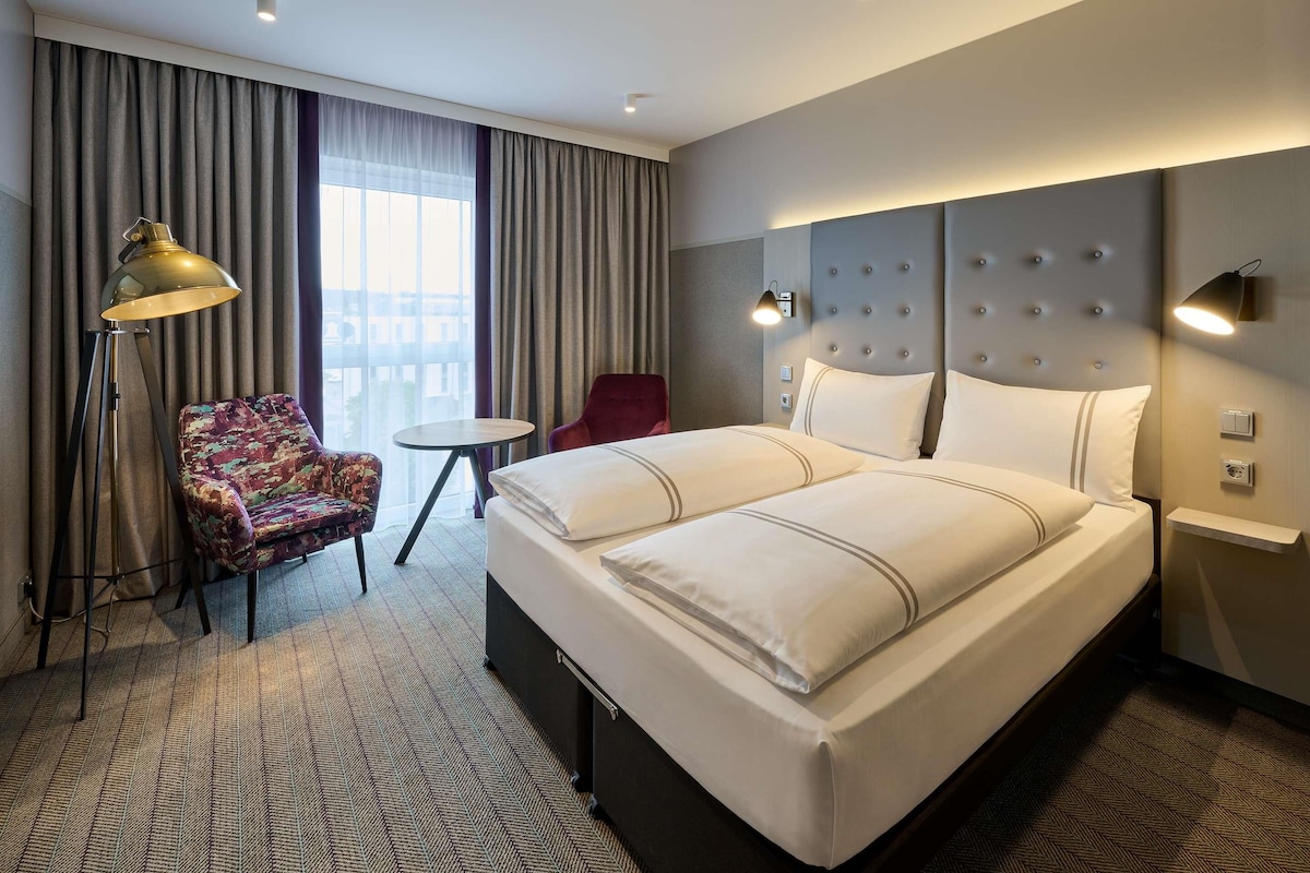 A hotel room with a large bed, a chair, a small table, and a lamp.