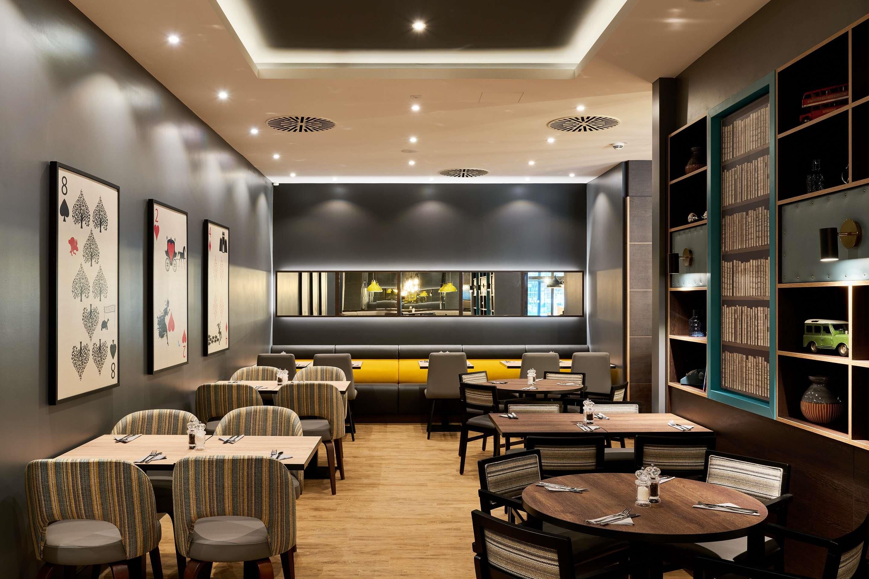Un comedor moderno con varias mesas y sillas, una zona de bar con mostrador, y una estantería llena de libros.