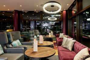 A modern lounge area with leather sofas, round wooden tables, and a bar visible through the glass.