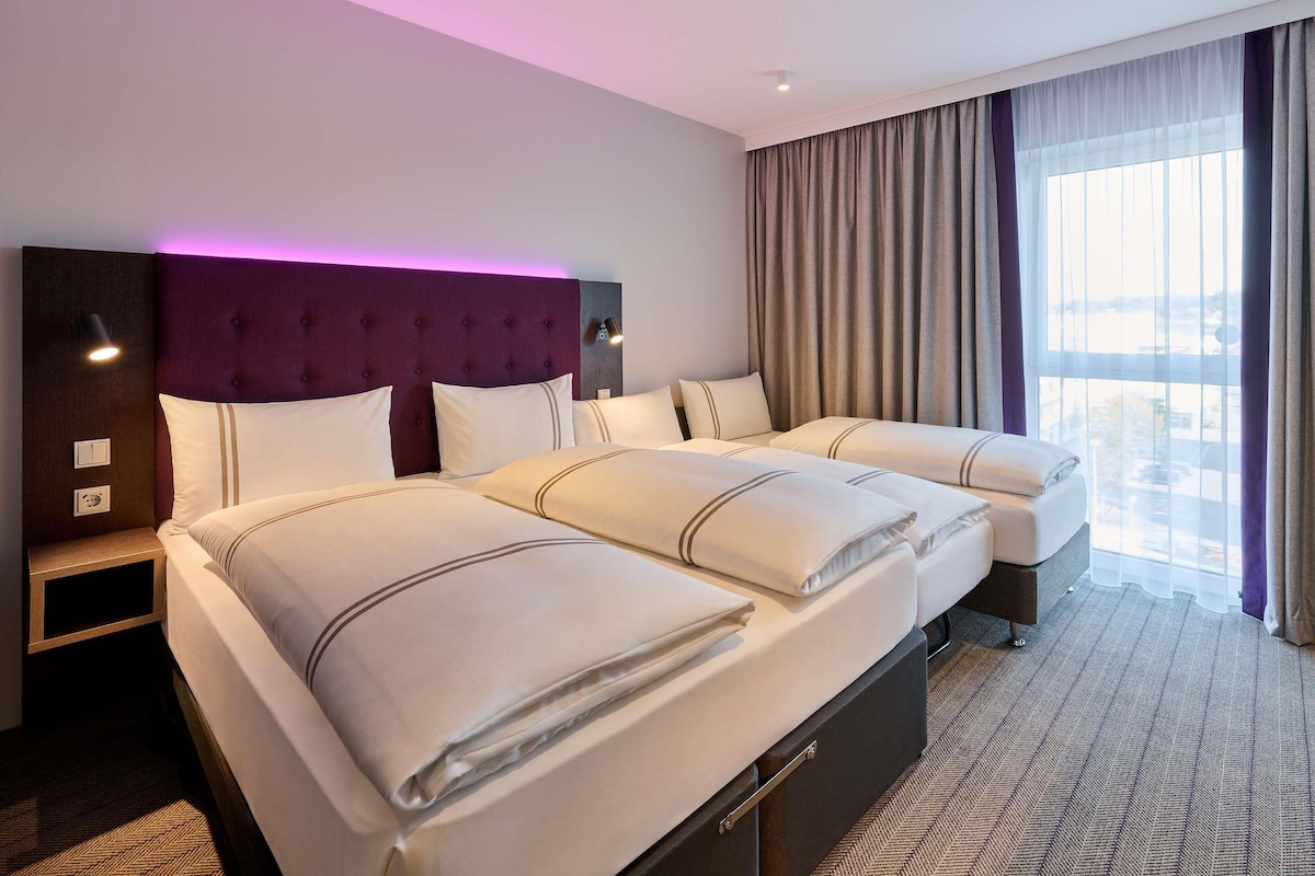 A hotel room with a large bed, a headboard, bedside lamps, a nightstand, and a view of the city through the window.