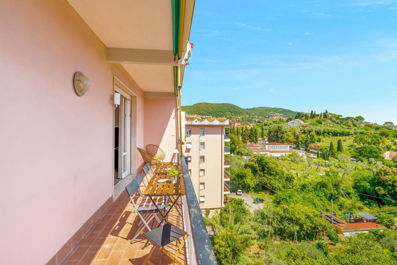 A balcony with a table and chairs, overlooking a green landscape with buildings.