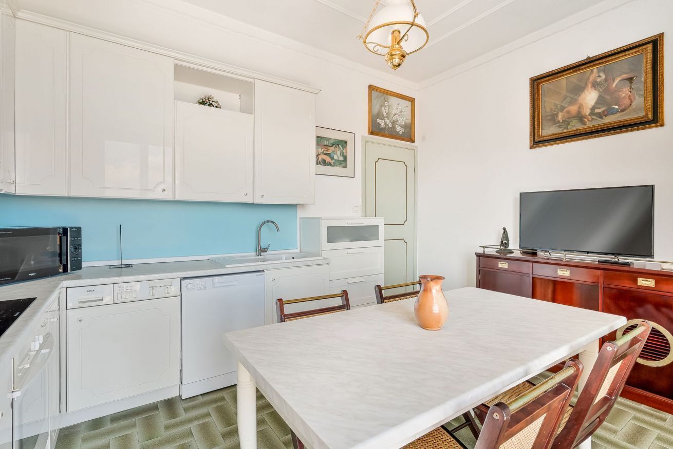Une cuisine compacte avec des meubles blancs, un four à micro-ondes, un lave-vaisselle et un évier. On y trouve une table à manger avec des chaises et un vase. Un téléviseur est posé sur un meuble en bois.
