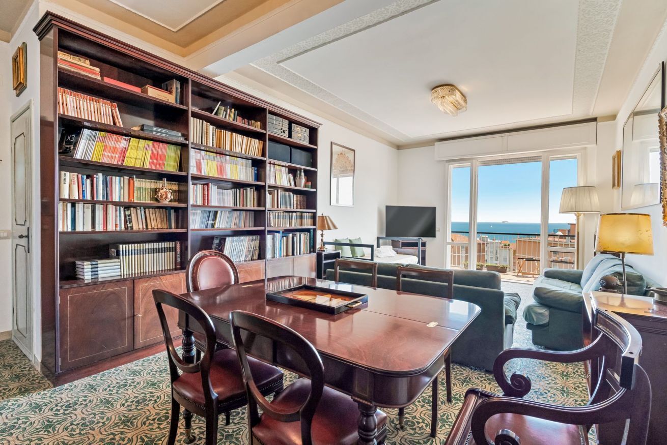 Un soggiorno spazioso con una grande libreria, un'area pranzo con tavolo e sedie, e vista sul mare attraverso ampie finestre.