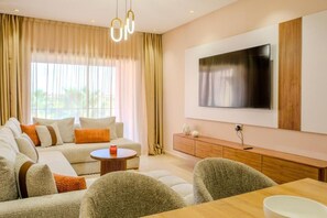 A modern living room with a sectional sofa, a dining table, and a flat-screen TV mounted on the wall. - Luxury 2BD in Prestigia Jade with Pool and Golf View (Marrakech)