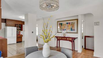 A dining area with a round table, chairs, a vase with yellow flowers, a chandelier, and a kitchen in the background.