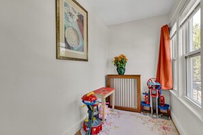 A corner room with a framed picture, a radiator, a small table with a toy, and a window with curtains.