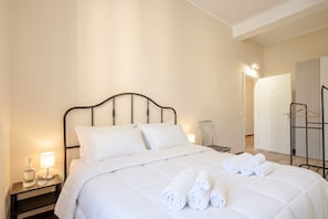 A neatly made bed with white linens and rolled towels, a bedside table with a lamp, and a wall-mounted chair. - ALL Covi del Viaggiatore – Elegant 2-Bedroom Stay in Bari (Bari)