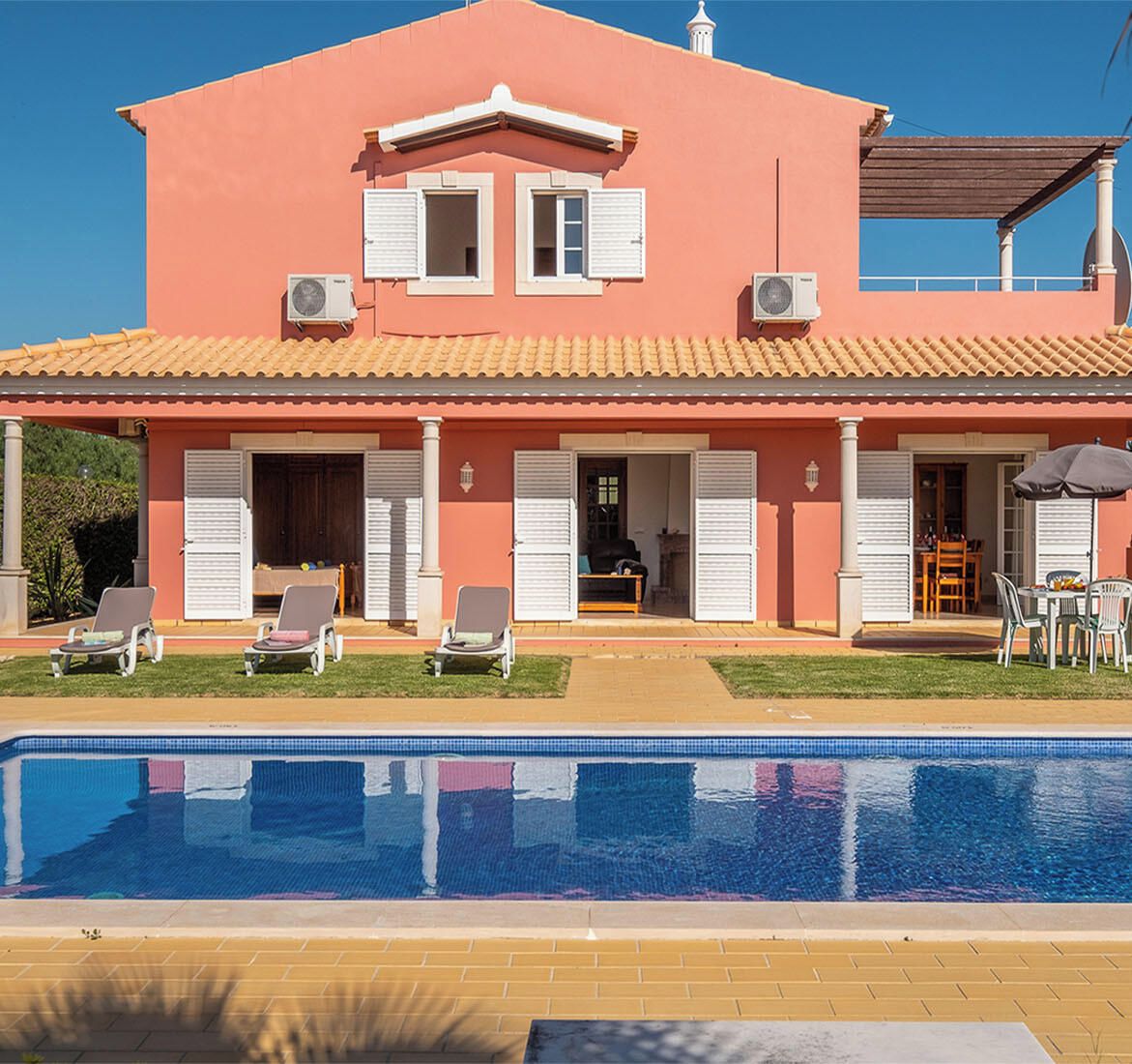 A two-story building with a swimming pool in front. There are lounge chairs and a table with umbrellas.