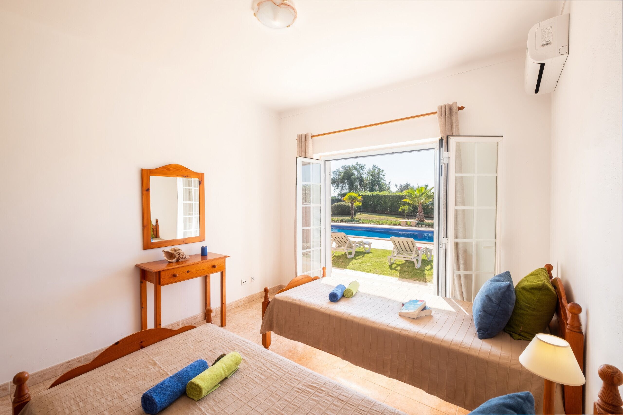 Une chambre avec un lit, une table de chevet, un miroir, une fenêtre donnant sur une piscine et un espace verdoyant, et un ventilateur de plafond.