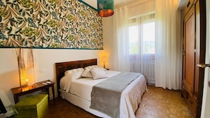 Ein Schlafzimmer mit Bett, Nachttisch, Lampe und Blick ins Freie durch ein Fenster.