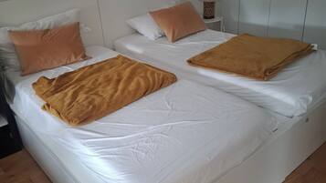 A double bed with white bedding and two brown blankets, a bedside table with a lamp, and a white wall.