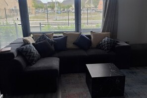 A sectional sofa with multiple cushions, a marble coffee table, and a view through a window with blinds. - Modern 2BDR Close to Dublin Airport and Coast (Lusk)