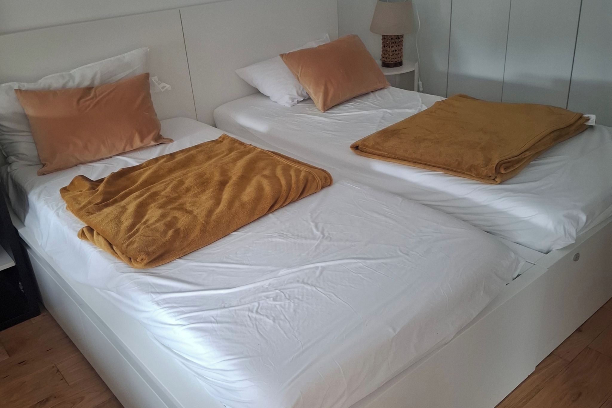 A double bed with white bedding and two brown blankets, a bedside table with a lamp, and a white wall.
