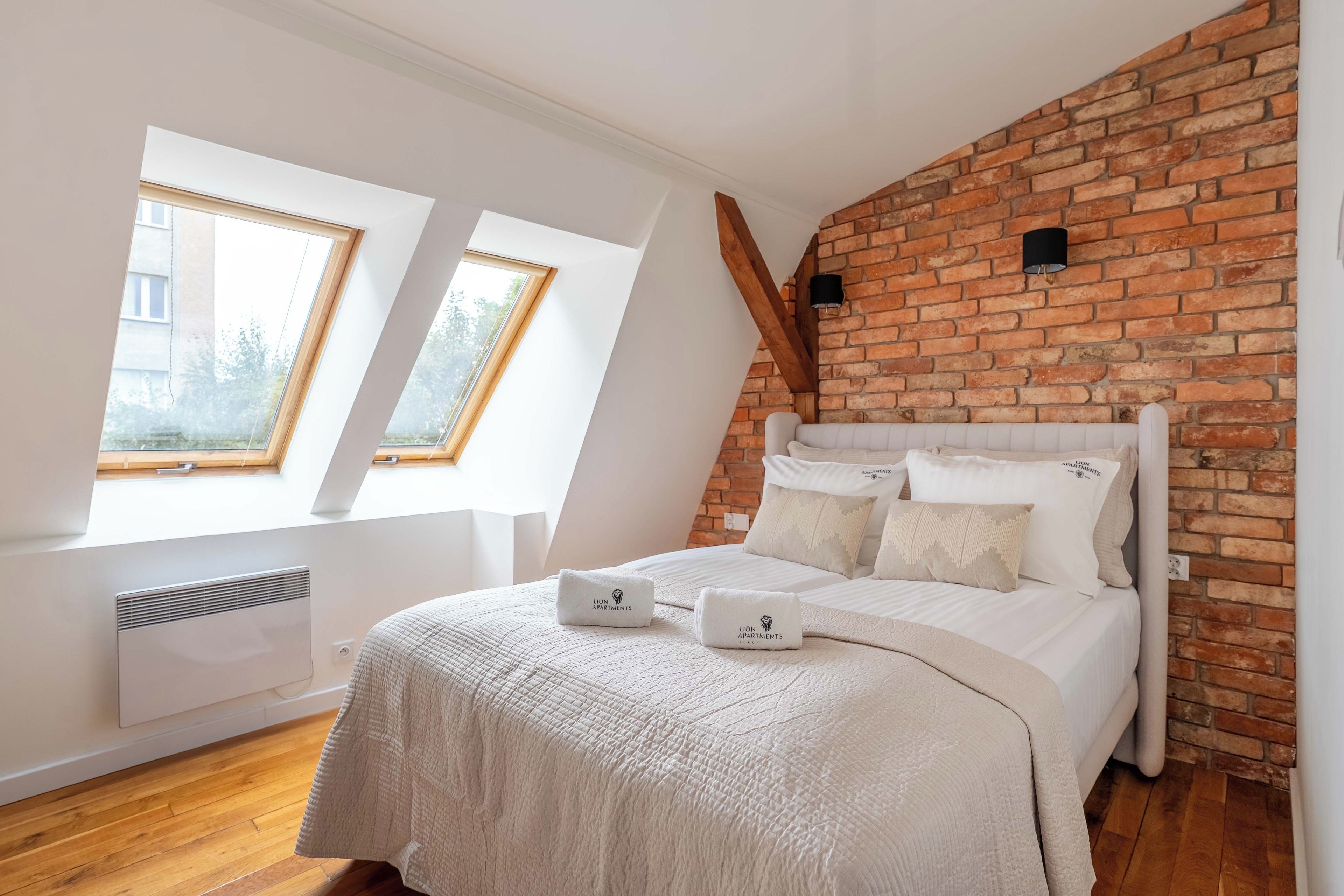 A bedroom with a sloped ceiling, a brick wall, a bed with white bedding, and two windows.