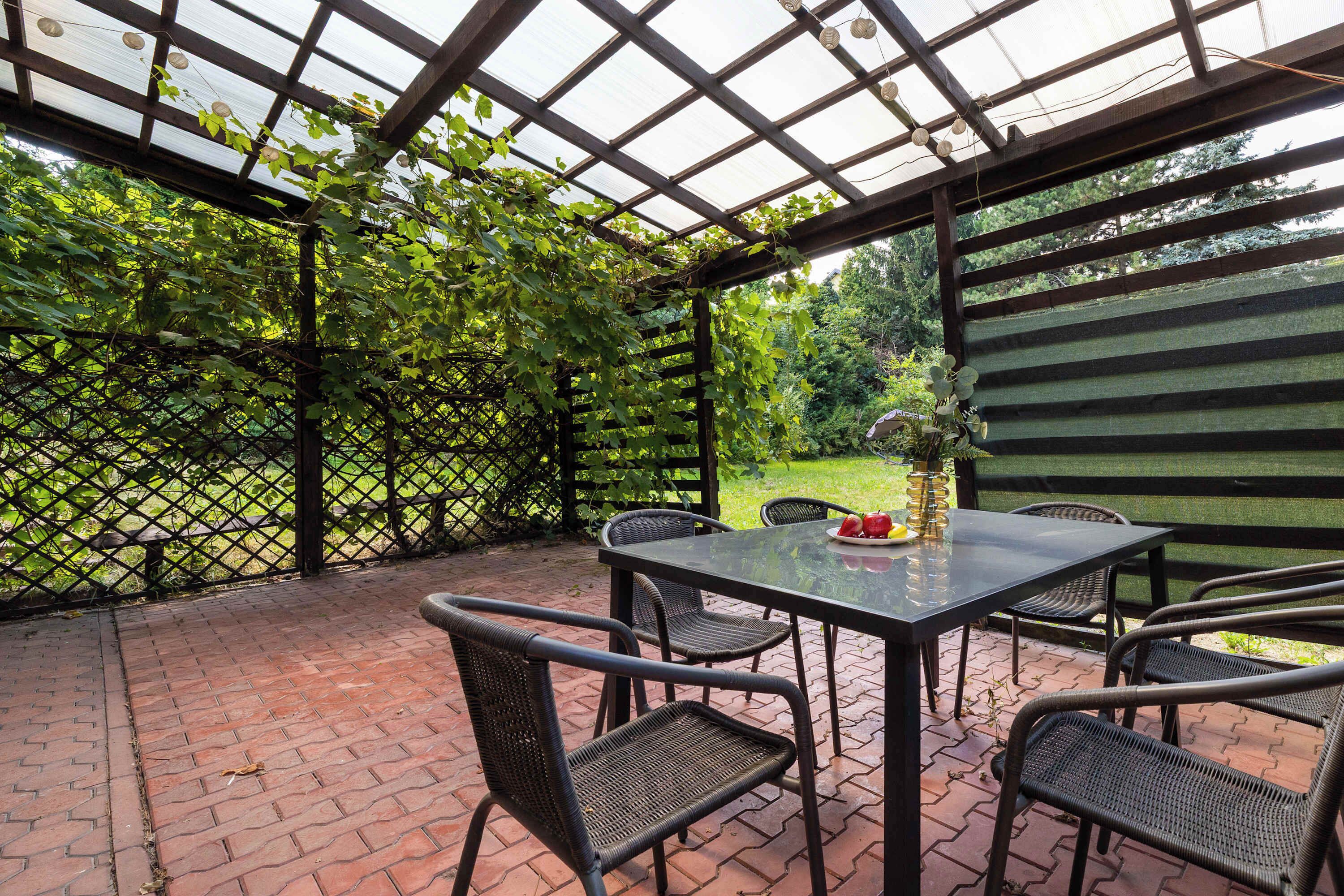 Eine Terrasse im Freien mit Tisch und Stühlen, umgeben von einer Holz-Pergola und einem Gitterzaun.
