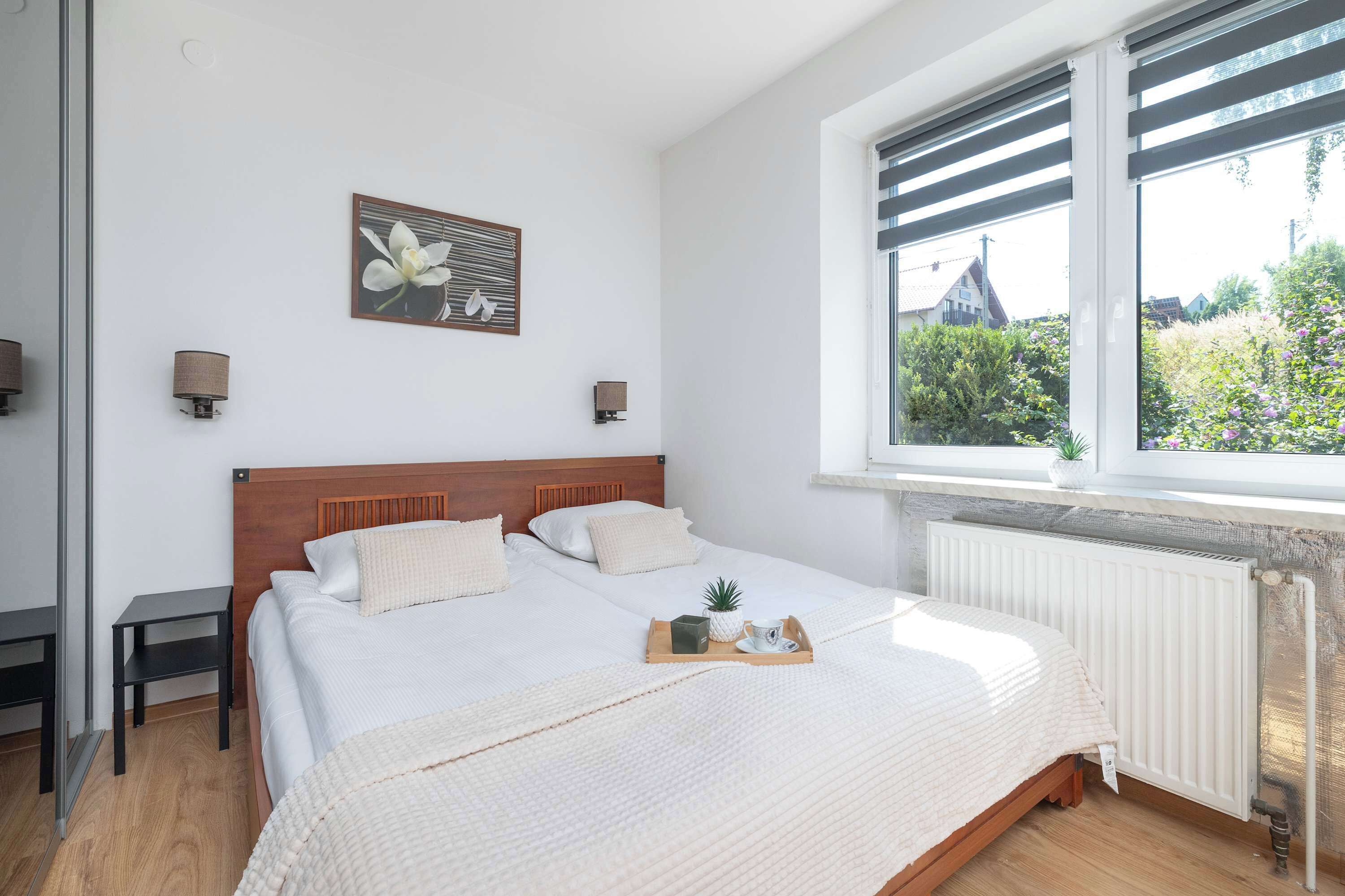 Ein Schlafzimmer mit hölzernem Kopfteil, zwei Nachttischen, einem Bett mit weißer Bettwäsche, einem Fenster mit Jalousien und Blick ins Grüne.