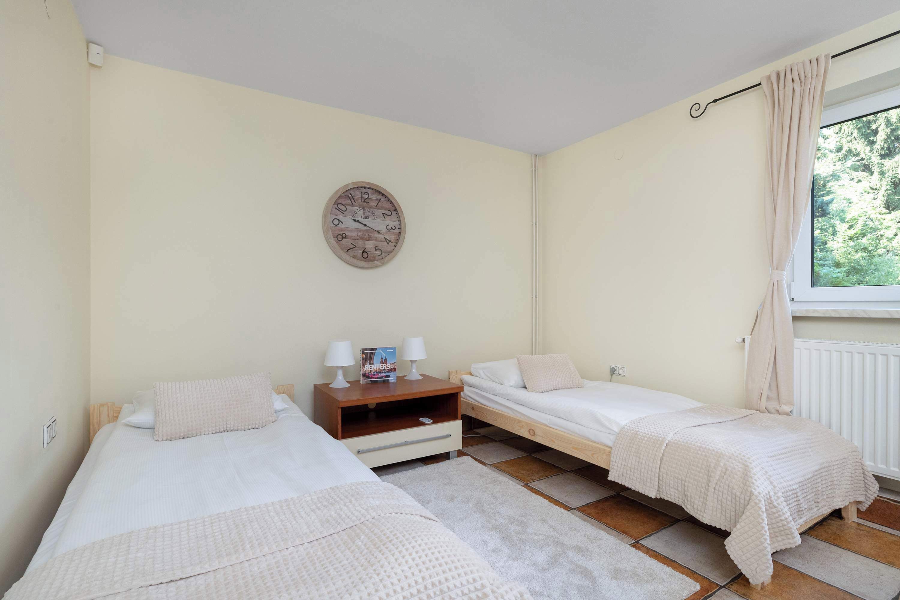 Ein Schlafzimmer mit zwei Einzelbetten, einem Holznachttisch, einer Wanduhr und einem Fenster mit Vorhängen.