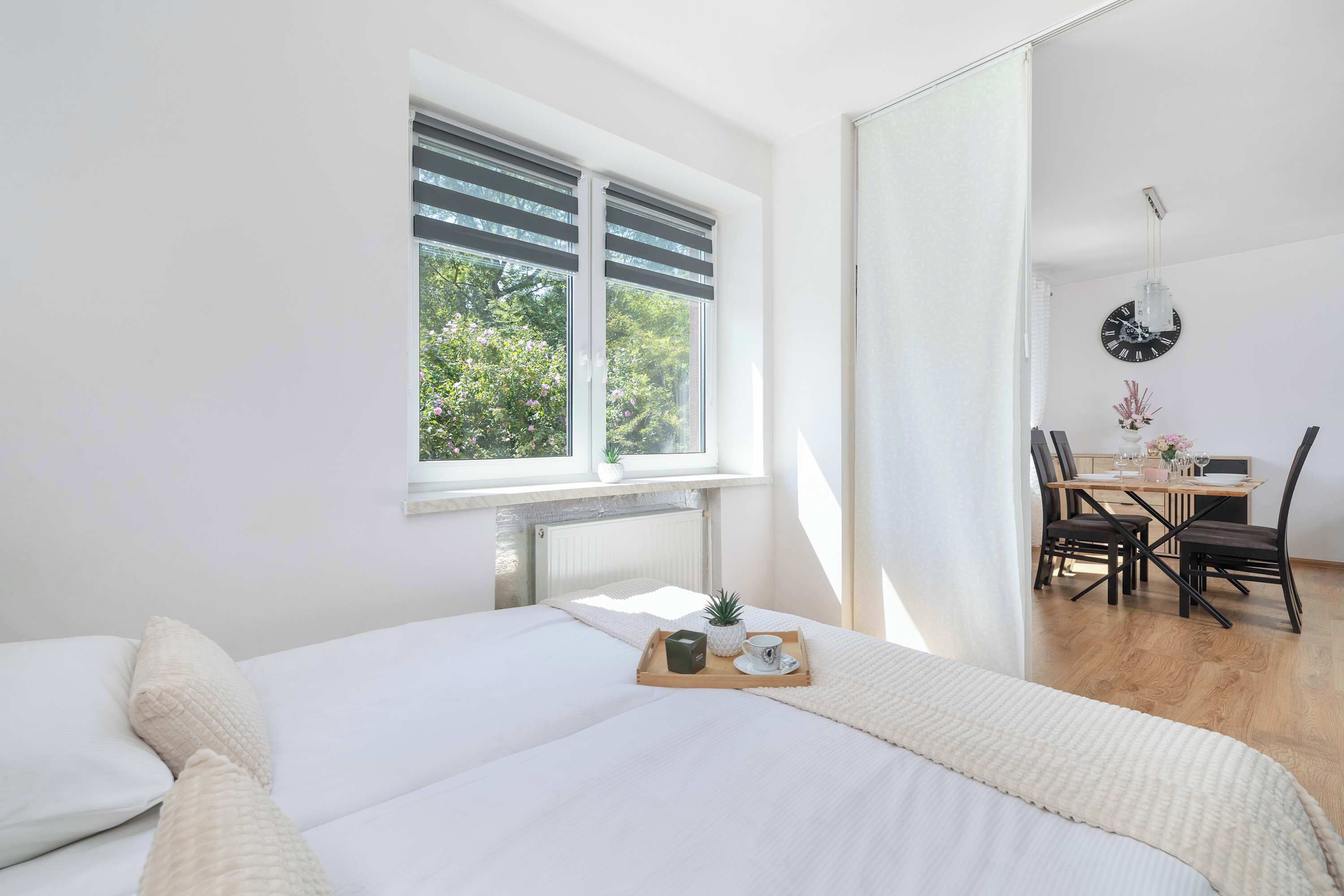 Une chambre avec une grande fenêtre, un lit recouvert de linge de lit blanc, une table de chevet avec un plateau, et un coin repas avec une table et des chaises.