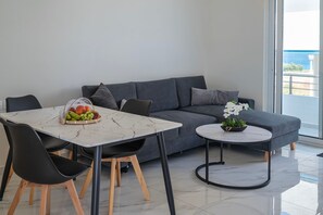 A modern living room with a grey sofa, a dining table with chairs, a small round table with a plant, and a view of the sea through a large window. - Kamiros Seaside Apartment in Kamiros Skala (Rhodes)