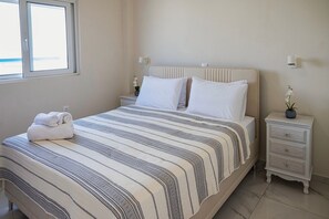 A neatly made bed with a striped bedspread, a bedside table with a lamp, and a window with blinds. - Kamiros Seaside Apartment in Kamiros Skala (Rhodes)