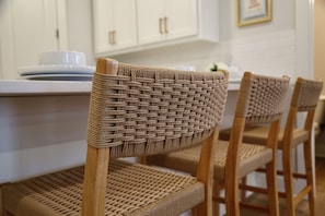 Wicker-Sessel mit Holzbeinen und Geflecht-Lehne stehen um eine Kücheninsel mit weißen Schränken und einer weißen Arbeitsplatte.