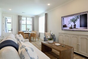 Een woonkamer met een bank, een salontafel, een eettafel met stoelen, een aan de muur bevestigde televisie, en een raam met gordijnen.