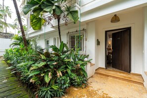 A building with a dark wooden door, a window with metal bars, and a balcony with lush green plants. - Staymaster Divli - 3BR Villa in Candolim (Candolim)