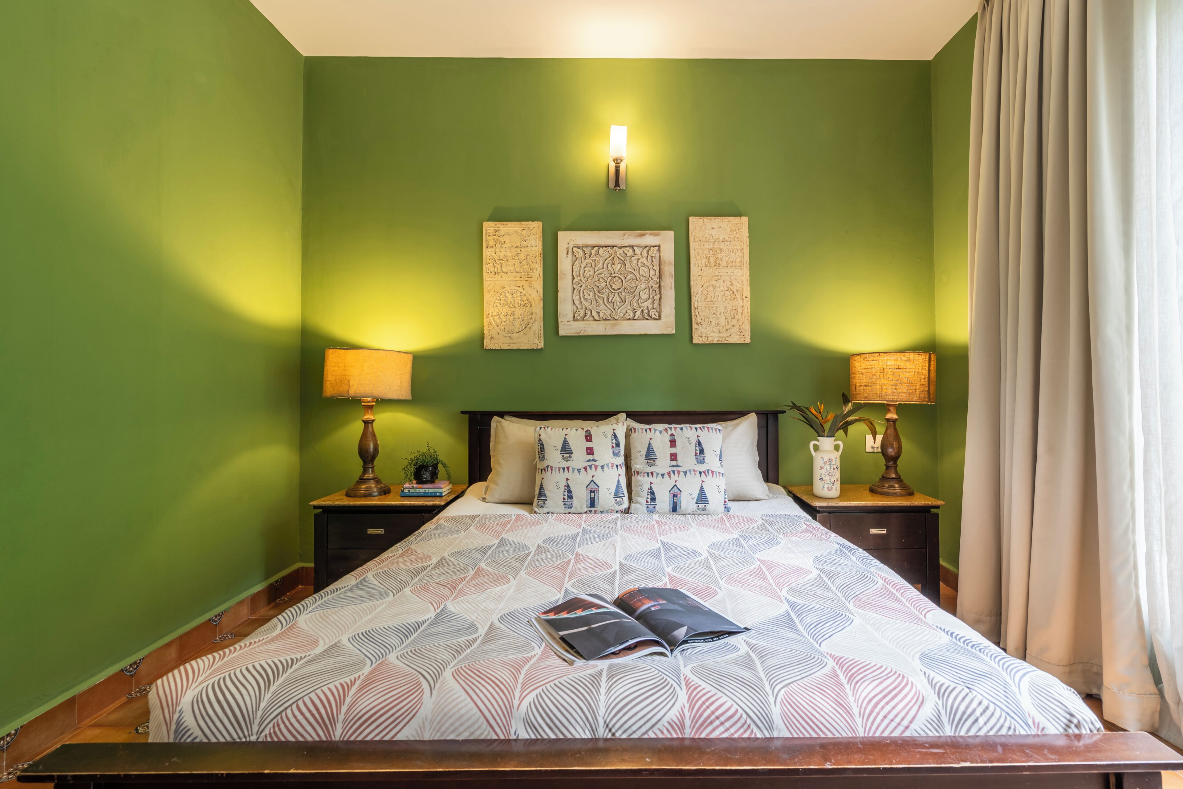 A bedroom with a bed, two bedside tables, a nightstand, a wall-mounted lamp, and a window with curtains.