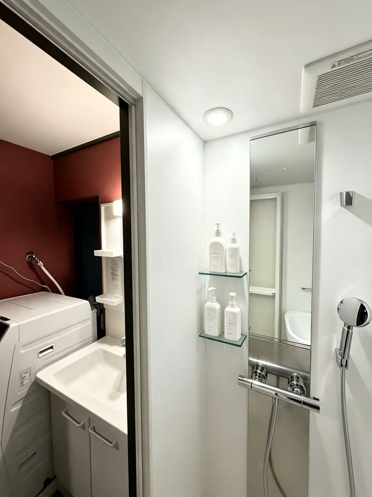 A modern bathroom with a built-in washing machine, a glass shelf with toiletries, and a mirror with a showerhead.